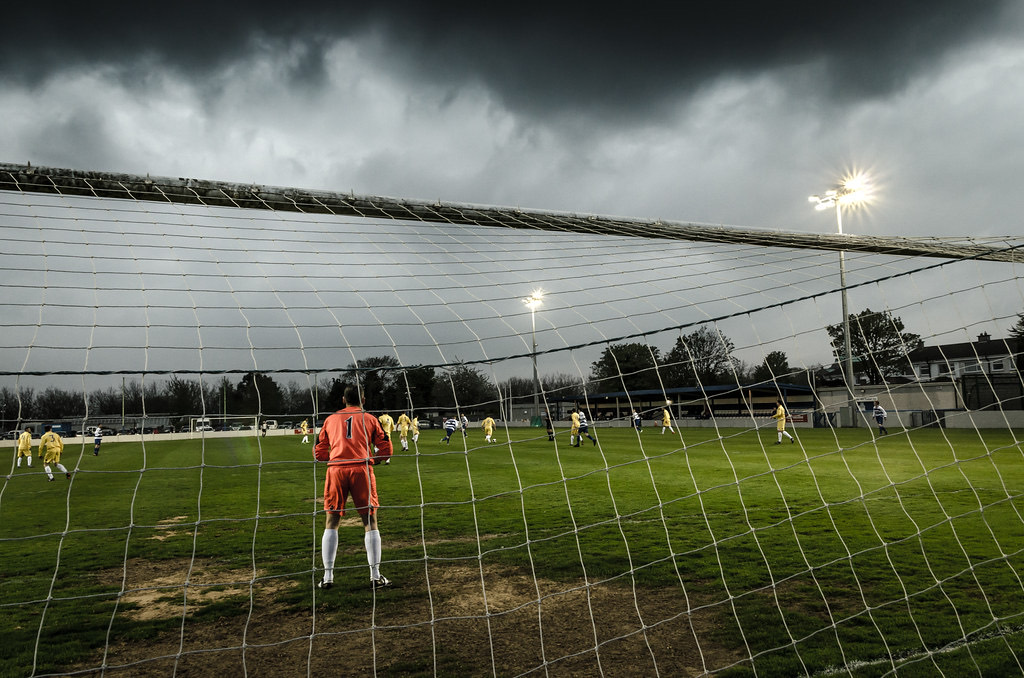 A goalkeeper seen through the net under floodlights and a storm sky