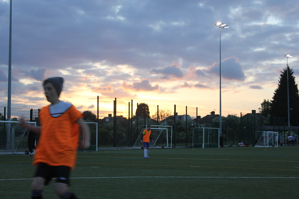 Players in orange bibs on an artificial pitch as the sun goes down